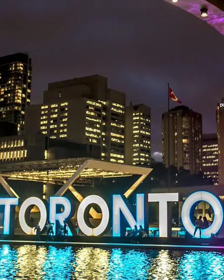 Menschen genießen den Abend vor und um das große Toronto-Schild im Freien, das in mehreren Farben beleuchtet ist und als Mittelpunkt des von hohen Gebäuden umgebenen Rathauses dient.