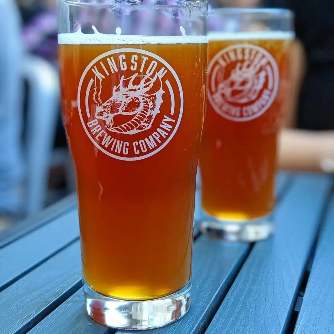 Two glasses of beer resting on a table.  One glass is close up in the foreground, one glass is in the background. 