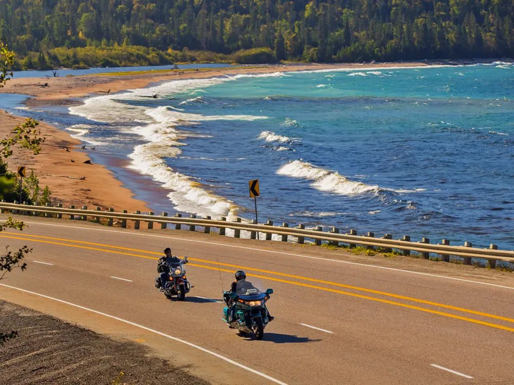 Two motorcyclists ride past a sandy beach with white capped waves crashing in.