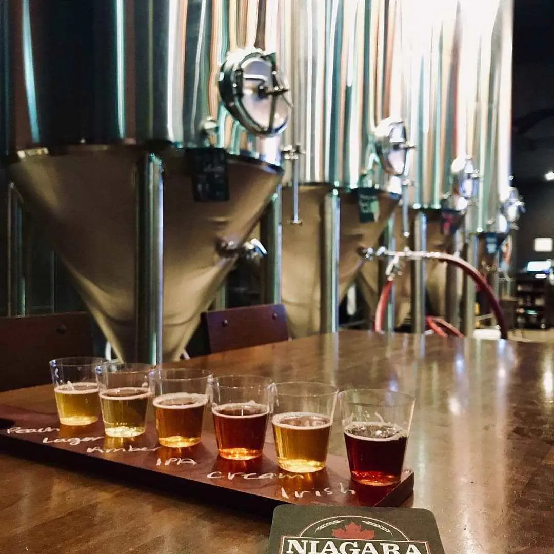 Un vuelo de seis cervezas de muestra se sienta en una mesa en primer plano, mientras que el equipo de elaboración se muestra en el fondo.