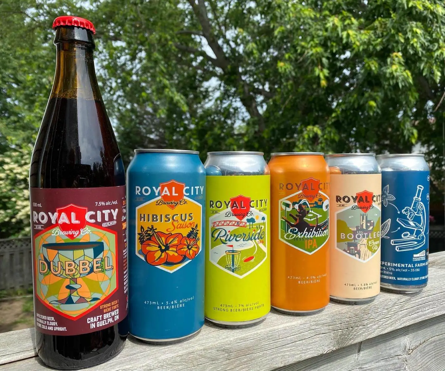 A tall dark bottle of beer beside 5 cans of beer resting on a fence