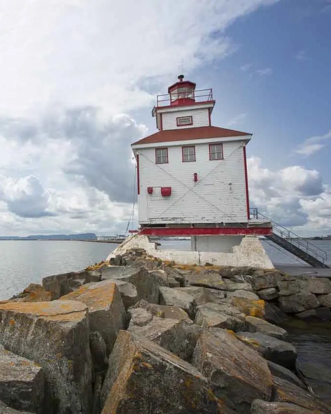 A sail boat sails past a white and red lighthouse atop a rocky coastline