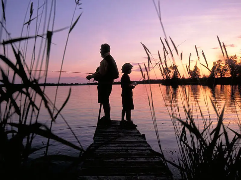 During the morning sunrise, a father and son fish back to back on a dock.