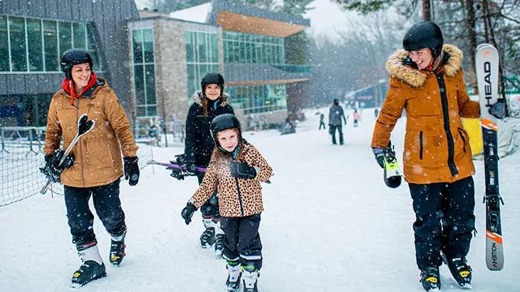 Two smiling adults and two children walk across snow in ski boots while holding skis.