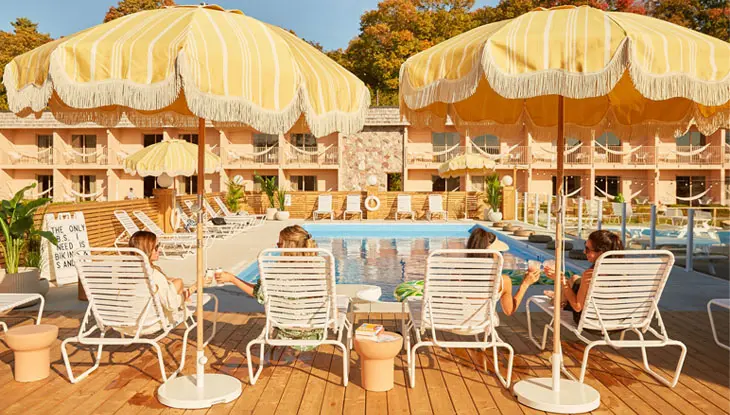 Quatre femmes relaxant sur des chaises longues au bord d’une piscine, sous des parasols jaunes.