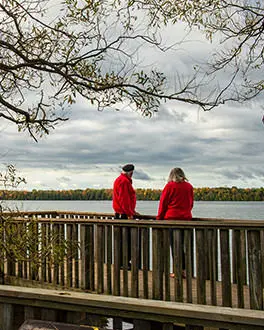A couple standing on a boardwalk, under a tree looking out at the lake. The trees on the shore in the distance are a mix of green and yellow.