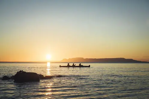 夕日を背景に眠っている巨人の姿が浮かび上がる中、3人がカヌーで湖を渡っています。