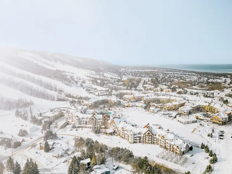 Aerial view of a snow-covered ski village with chalet-style buildings, tree-lined runs, and Georgian Bay visible in the distance.