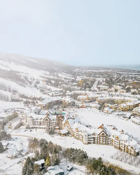 Aerial view of a snow-covered ski village with chalet-style buildings, tree-lined runs, and Georgian Bay visible in the distance.