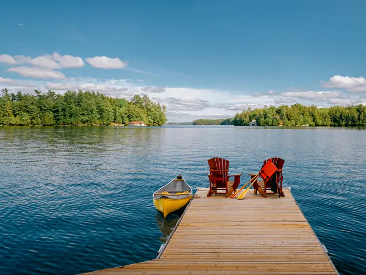 A wooden dock with Adirondack chairs and a yellow canoe, overlooking a calm lake and forested shoreline under a clear blue sky