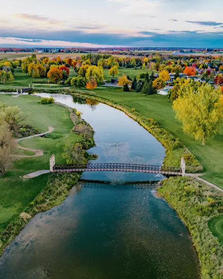 A vibrant fall scene of a river winding through a golf course, with a pedestrian bridge and a distant town on the horizon.