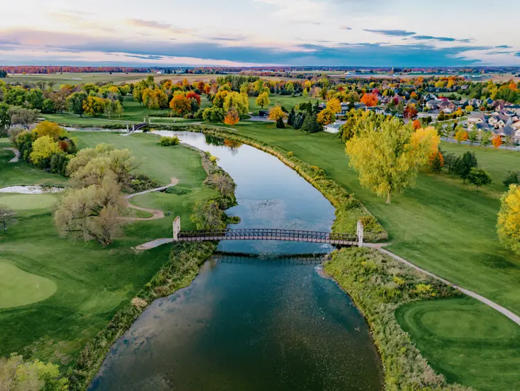 A vibrant fall scene of a river winding through a golf course, with a pedestrian bridge and a distant town on the horizon.
