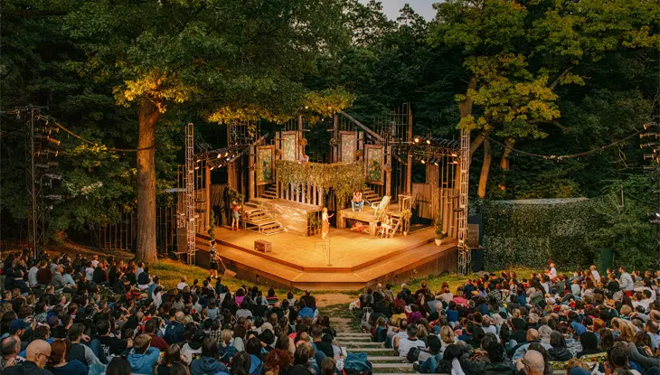 황혼 무렵, 나무에 둘러싸인 야외 원형극장에 군중이 모여 따뜻하게 밝혀진 무대에서 연극이 펼쳐지는 것을 지켜보았습니다.