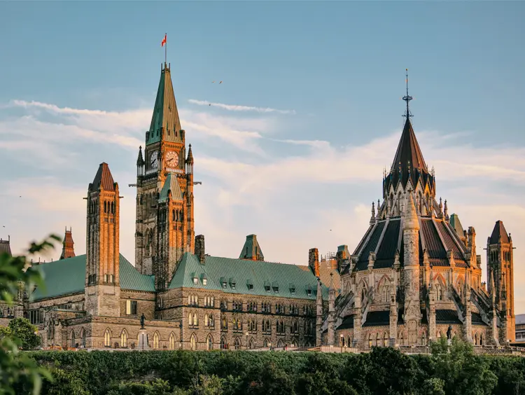 Looking out from Major’s Hill Park toward iconic gothic towers glowing in sunset light, framed by greenery and a soft blue sky.