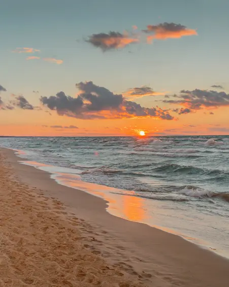 A sandy beach along Lake Huron at sunset, with the sun reflecting on the water and waves gently rolling ashore under a colourful sky.