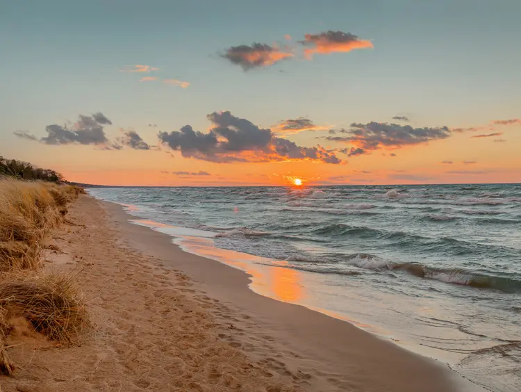 A sandy beach along Lake Huron at sunset, with the sun reflecting on the water and waves gently rolling ashore under a colourful sky.