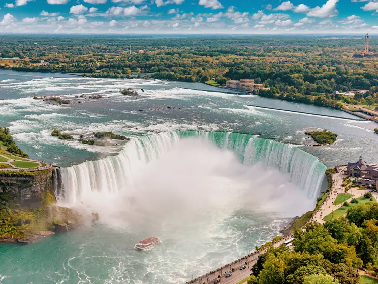 Aerial view of Horseshoe Falls on the Canadian side, with a sightseeing boat below and mist rising under a blue sky.