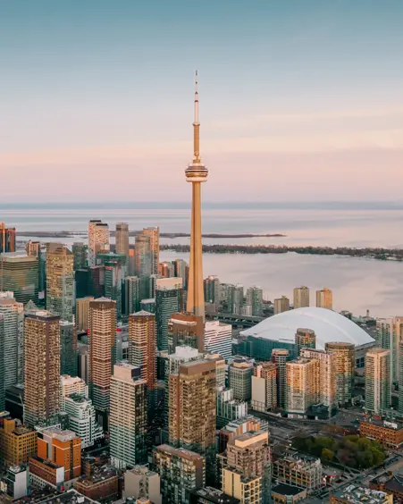Skyscrapers and the CN Tower bathed in sunset light, overlooking calm waters under a soft, pastel sky.
