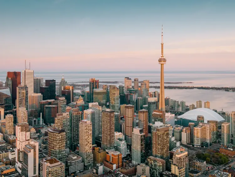 Skyscrapers and the CN Tower bathed in sunset light, overlooking calm waters under a soft, pastel sky.