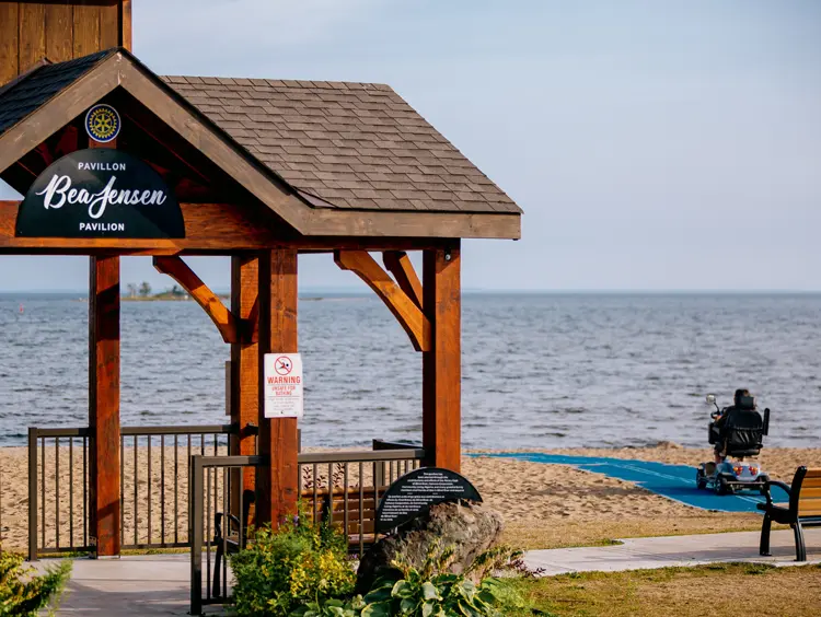 Pabellón de madera con vista a la playa y al agua en Blind River, con un scooter de movilidad en un camino accesible que conduce a la costa.