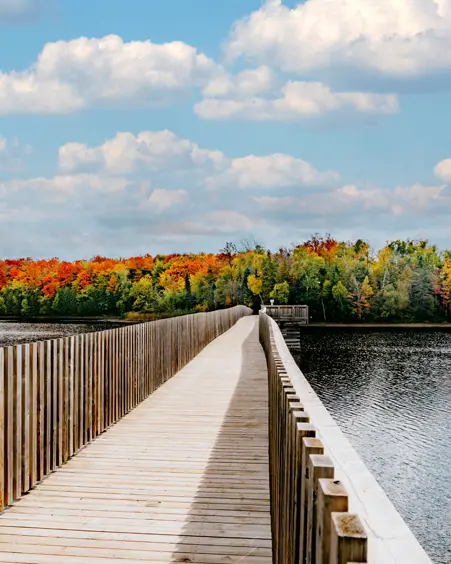 Strandpromenade in Orangeville, Ontario, die sich über den Island Lake in Richtung baumbedecktes Land erstreckt, mit leuchtenden Herbstfarben unter blauem, bewölktem Himmel.