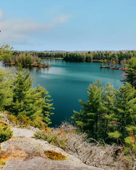 Blick auf den See, eingerahmt von felsigem Gelände und dichtem Kiefernwald im Frontenac Provincial Park an einem teilweise bewölkten Frühlingstag.