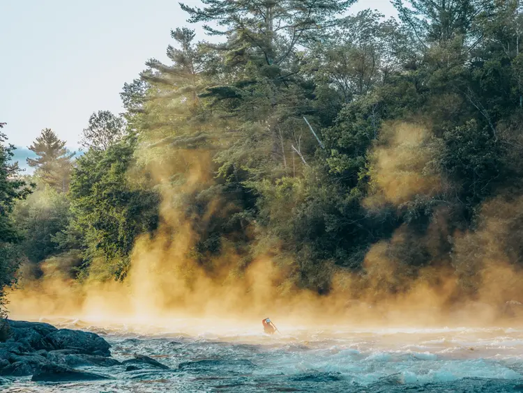 Paddler navigating misty rapids on the Madawaska River in the Haliburton Highlands, surrounded by dense forest and early morning light.