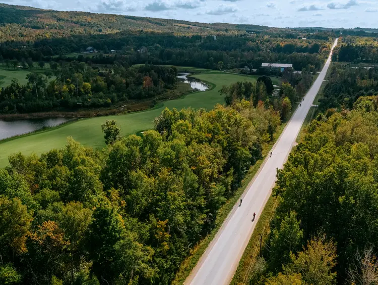 Vista aérea de ciclistas recorriendo un camino rural en Thornbury, bordeado por un bosque y un río sinuoso en un día de verano parcialmente nublado.