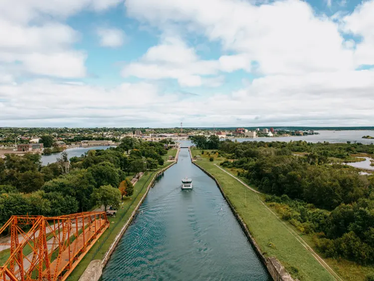 A boat cruising through the Sault Ste. Marie Canal on a cloudy summer day, passing beneath a red swing bridge with greenery along the shoreline.