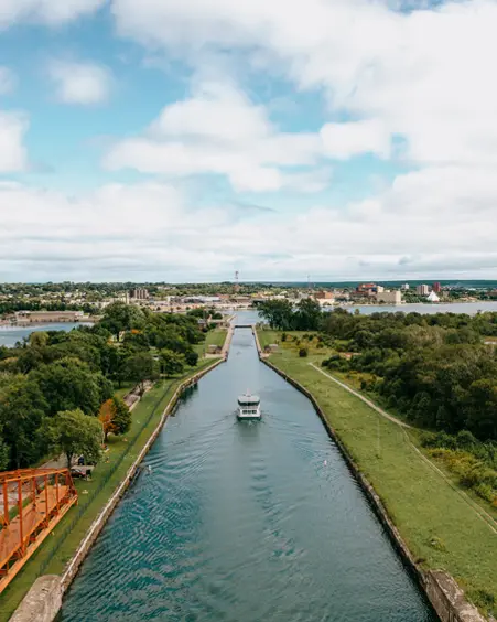 Ein Boot fährt an einem bewölkten Sommertag durch den Sault Ste. Marie Canal und passiert dabei eine rote Drehbrücke mit viel Grün entlang der Küste.