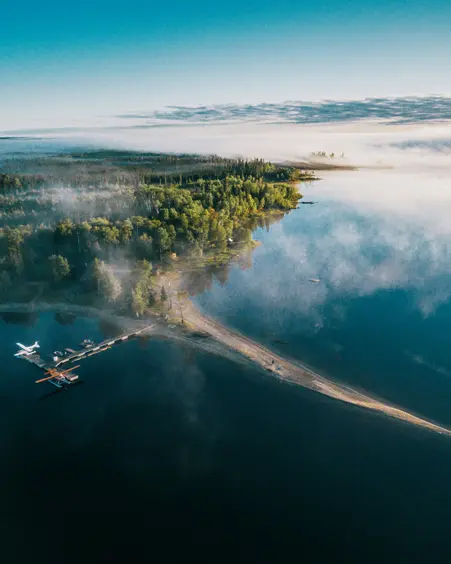  Vista aérea del Parque Provincial Wabakimi, con un hidroavión atracado, un denso bosque verde y niebla flotando sobre un lago quieto y cristalino al amanecer.