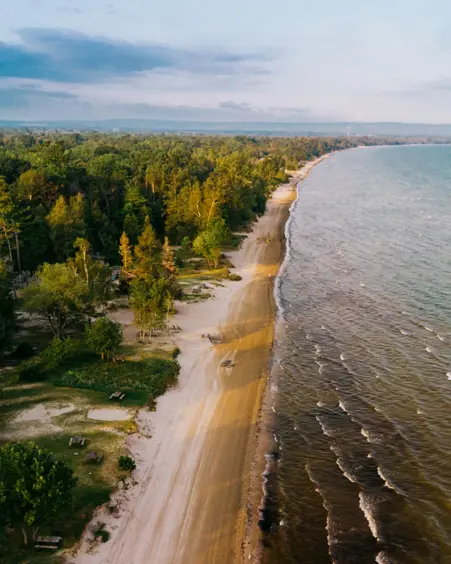Aerial view of Wasaga Beach with a wide sandy shoreline, forested parkland bordering the beach, and gentle waves under a pastel evening sky.