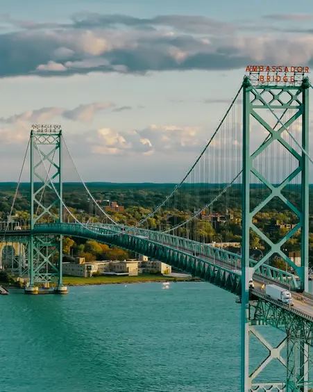 Aerial view of the Ambassador Bridge stretching across turquoise waters, connecting Windsor, Ontario, with the U.S., under a sunset sky.