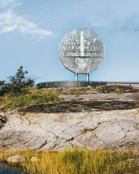 A giant silver-coloured replica of a nickel coin, the Big Nickel, standing above rocky terrain and a pond at Dynamic Earth, Sudbury, on a summer day.