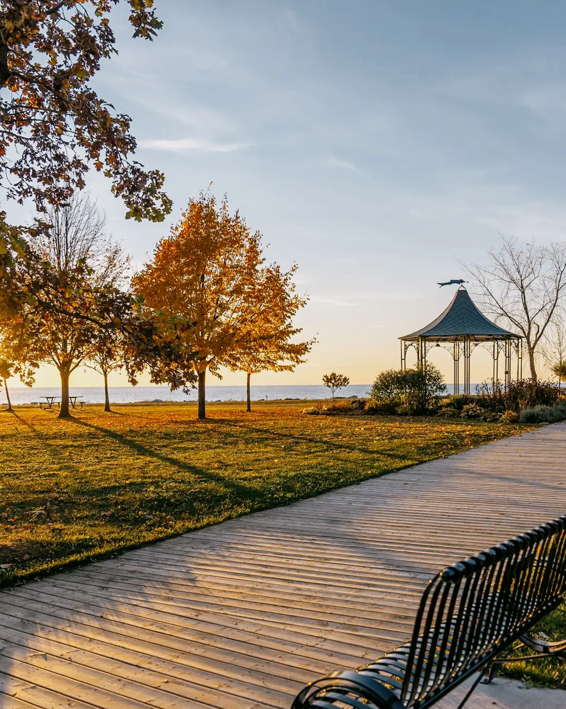  Ein von Bäumen und einer schwarzen Bank gesäumter Holzsteg, der bei Sonnenuntergang zu einem Pavillon an der Uferpromenade von Goderich führt.
