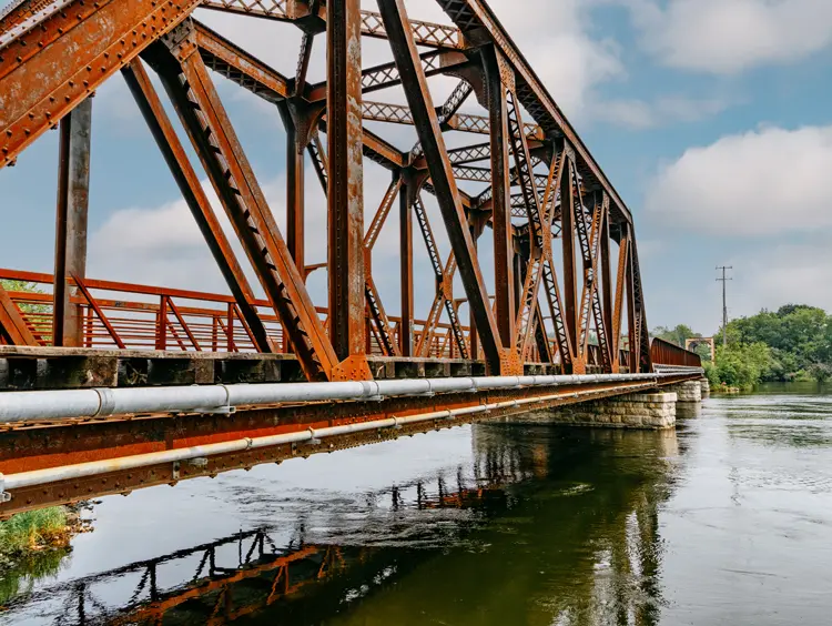 Puente ferroviario de color óxido sobre el río Otonabee a lo largo del Sendero Transcanadiense en Peterborough, reflejado en aguas tranquilas.