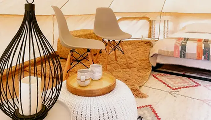 Deux chaises beiges, un pouf en tricot et un plateau contenant deux tasses, un lit et un bougeoir décoratif à l’intérieur d’une tente, à Lakecroft.