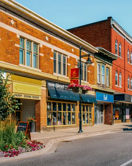Colourful historic storefronts in downtown Parry Sound, with bright flower beds along the sidewalk and hanging baskets on a clear summer day.
