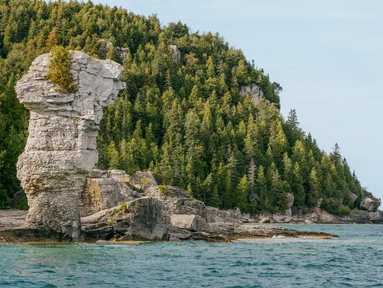 A massive flowerpot rock formation rising from the shoreline of Flowerpot Island in Tobermory, with a forested cliff in the background.