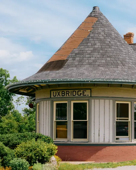  Der historische Bahnhof von Uxbridge mit einem runden Turmdach, cremefarbener Außenverkleidung und großen Fenstern, umgeben von Pflanzgefäßen an einem Sommertag.