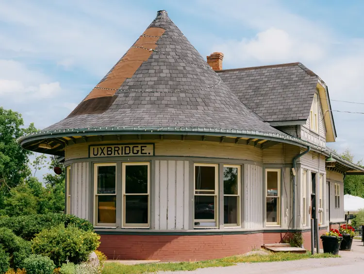  Der historische Bahnhof von Uxbridge mit einem runden Turmdach, cremefarbener Außenverkleidung und großen Fenstern, umgeben von Pflanzgefäßen an einem Sommertag.