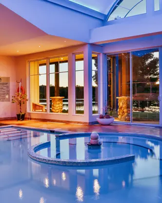 オンタリオ州イーグル レイクにある Sir Sam's Inn & Spa の青い照明、大きな窓、水面に映る夕日の眺めが楽しめるモダンな屋内プール。