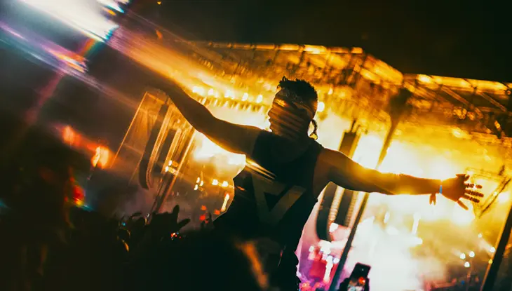 Participant au festival VELD assis sur les épaules d’une autre personne, les bras tendus, tandis que les lumières vives de la scène les éclairent.