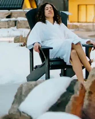 Femme en robe de chambre blanche relaxant dans un fauteuil Muskoka au Vettä Nordic Spa en hiver, la vapeur s’élevant d’une piscine à proximité.