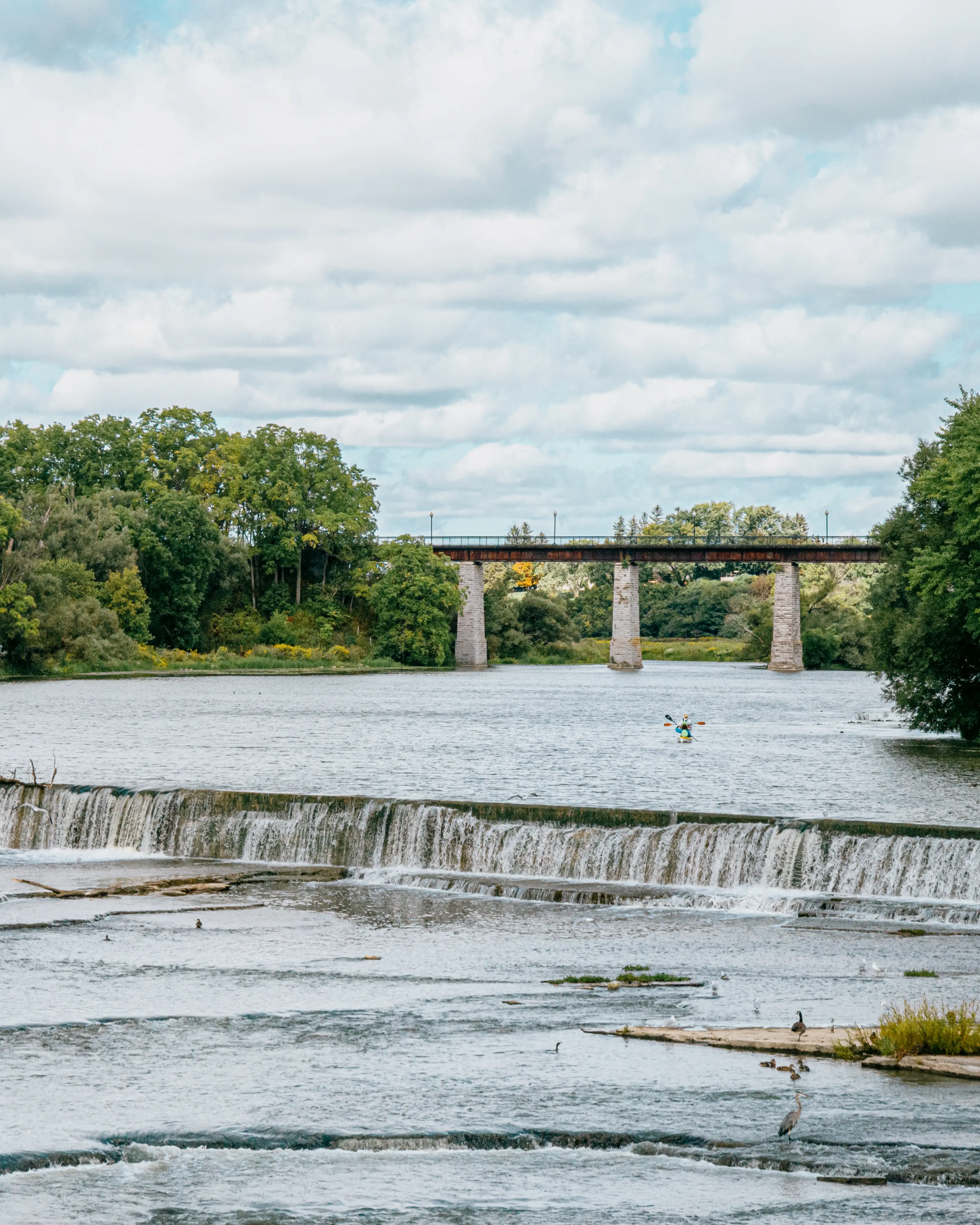  An einem teilweise bewölkten Sommertag stürzt Wasser über einen niedrigen Damm in St. Marys, Ontario, eingerahmt von dichten Bäumen und einer Steinbrücke in der Ferne.
