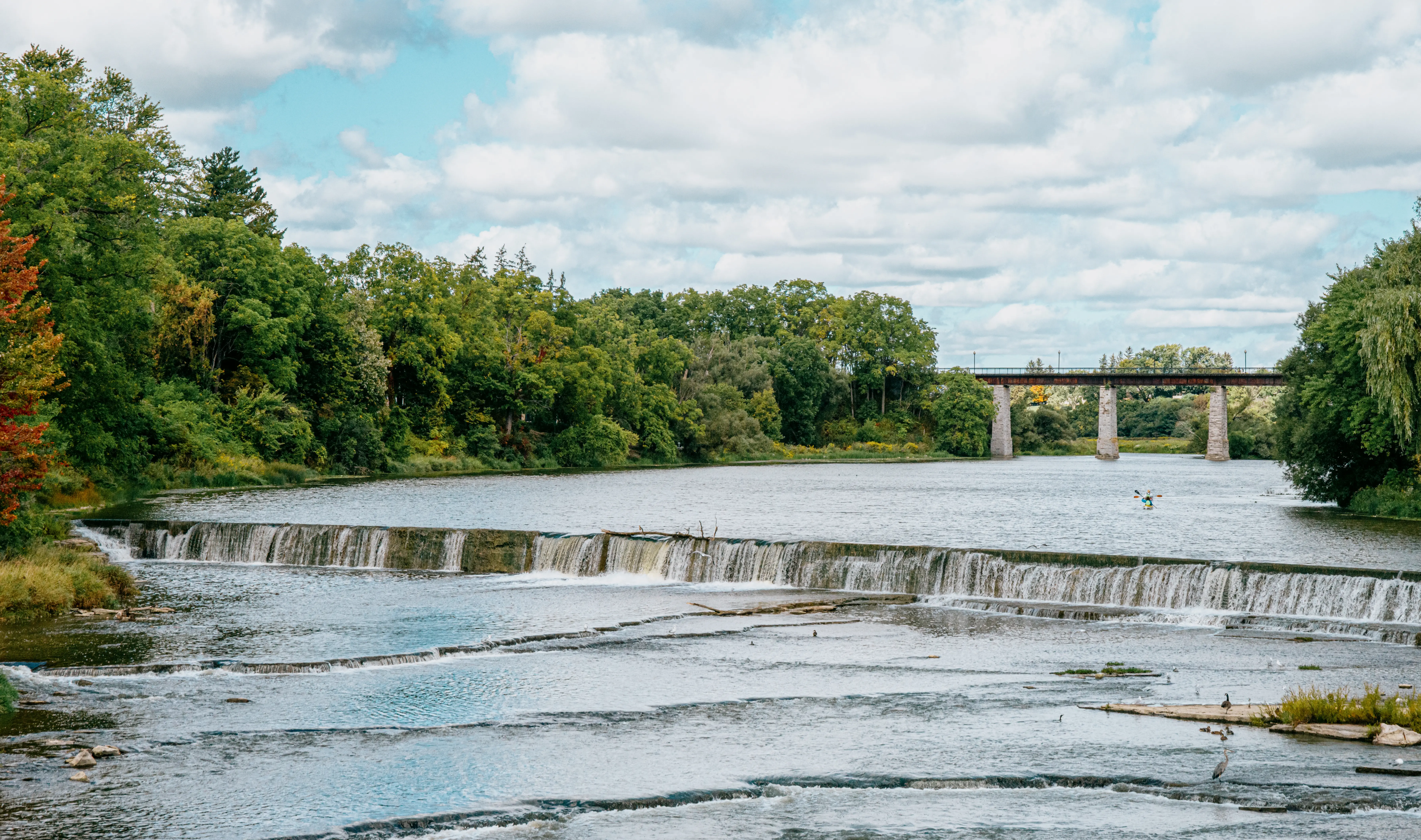  An einem teilweise bewölkten Sommertag stürzt Wasser über einen niedrigen Damm in St. Marys, Ontario, eingerahmt von dichten Bäumen und einer Steinbrücke in der Ferne.