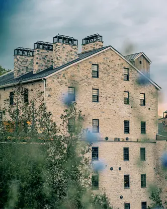 Bâtiment rustique en pierre de l’Elora Mill Hotel & Spa avec ses multiples cheminées, vu à travers des fleurs sauvages d’un bleu tendre au crépuscule.