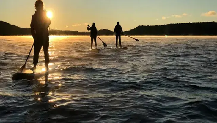 Trois visiteurs de Northern Edge Algonquin sur une planche à pagaie sur le lac Kawawaymog au coucher du soleil.