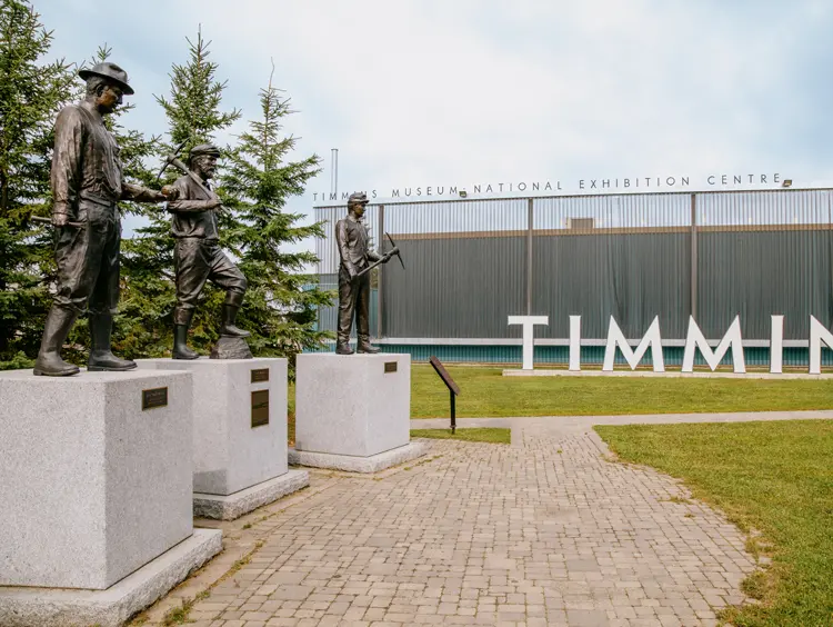 Estatuas de mineros afuera del Centro Nacional de Exposiciones del Museo Timmins, con grandes letras TIMMINS en el fondo en un día nublado.