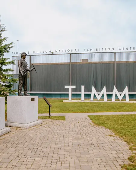 Estatuas de mineros afuera del Centro Nacional de Exposiciones del Museo Timmins, con grandes letras TIMMINS en el fondo en un día nublado.